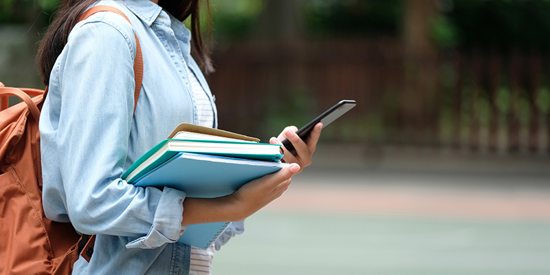 Female student holding books and a cell phone while wearing a backpack
